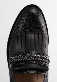 Mocassin en cuir noir avec texture crocodile, lanière à franges et détail en chaîne argentée sur le dessus, vu de dessus.