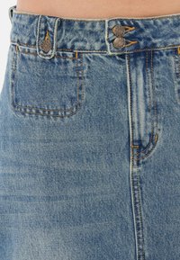 Lichtblauwe denimrok met een hoge taille, dubbele knoopsluiting, decoratieve stiksels en één achterzak. Vlotte textuur met een vervaagde afwerking.