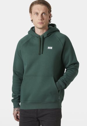 Jonge man met licht haar, gekleed in een donkergroene Helly Hansen hoodie, met hand in zijn zak, tegen een effen lichte achtergrond.