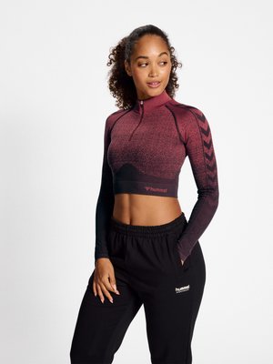 Kvinde med krøllet hår i en høj hestehale iført en bordeaux og sort langærmet cropped træningstop og sorte hummel træningsbukser med hænderne i lommerne.