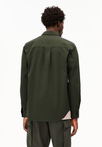 Man with short curly hair wearing a dark green long-sleeve shirt and cargo pants, facing away on a white background.