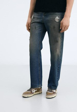 Jeans di denim blu sbiadito con vestibilità dritta, caratterizzati da dettagli strappati e leggeri segni di usura sulle cosce, abbinati a sneaker marroni.