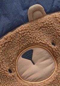 Pruun plush kangas tekstuuriga, koheva pinnaga. Omab beeži ümmargust plaastrit, millel on tikitud muster ja pehme, ümar kõrvakaunistus.