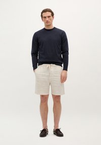 Granatowy sweter z dzianiny, jasnobeżowe lniane szorty z sznurkiem i ciemnobrązowe skórzane buty. Model stoi na jasnym tle.