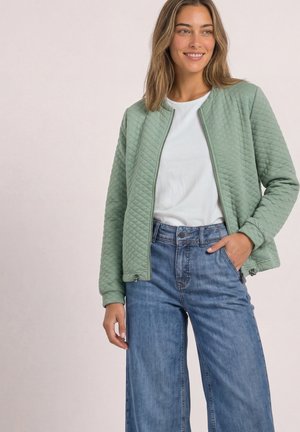 Frau trägt eine hellgrüne gesteppte Jacke über einem weißen Hemd und hoch taillierte blaue Jeans, steht mit einer Hand in der Tasche und lächelt.