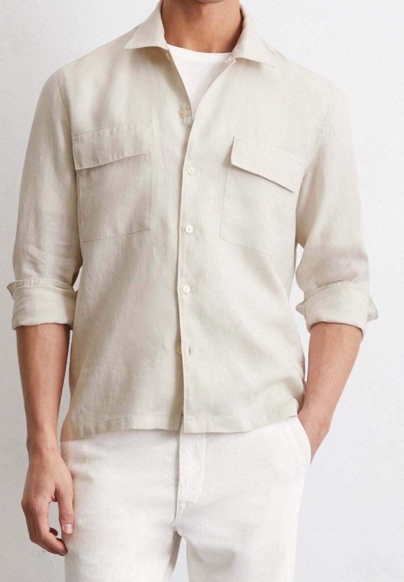 Homme portant une chemise beige clair boutonnée avec deux poches poitrine, manches retroussées et pantalon blanc, debout devant un fond blanc.
