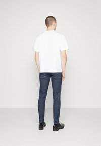 Vit t-shirt med korta ärmar, blå slim fit-jeans, svarta stövlar. Ren design med enfärgad textur, sedd bakifrån mot en neutral bakgrund.