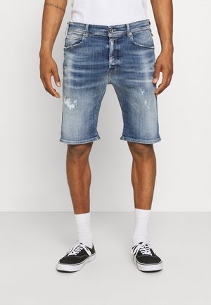 Mann in verblassten blauen Jeansshorts, weißen Kniestrümpfen und schwarzen Sneakers mit niedriger Schnittführung vor einem einfarbigen Hintergrund.