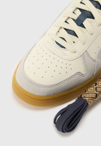 Skórzano-zamszowe sneakersy w kolorze kremowym i szarym, z perforacjami na czubku, ciemnoniebieskim językiem oraz gumową podeszwą z niebieskimi sznurówkami.