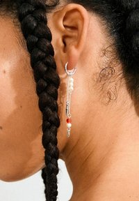 Silberner Creolenohrring mit hängenden Ketten; enthält weiße Perlen und eine rote Perle; vor einem Hintergrund aus geflochtenem Haar präsentiert.