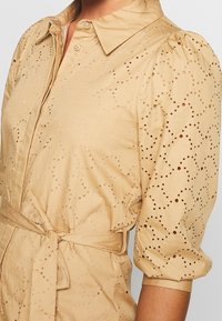 Vestido camisa en un tono beige claro con mangas abullonadas, presenta un cuello con solapa y un diseño perforado. Incluye un cinturón anudado en la cintura para dar forma.