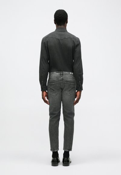 Camicia in denim nero con colletto a bottoni, indossata con jeans grigio scuro e scarpe nere, vista da dietro su uno sfondo chiaro.