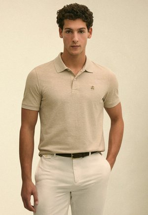 Jonge man met krullend haar, gekleed in een beige poloshirt dat in een witte broek is gestopt, staand tegen een effen lichte achtergrond.