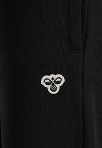 Schwarze Stoff-Jogginghosen mit einem kleinen, schwarz-weißen gestickten Bienenlogo auf der linken Seite. Glatte Textur, enganliegendes Design, elastischer Bund.