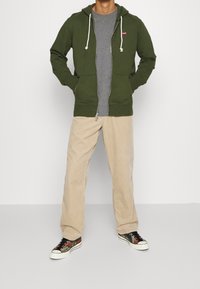 Felpa verde con zip e cordini sopra una maglietta grigia, abbinata a pantaloni in corduroy beige e sneakers camouflage.