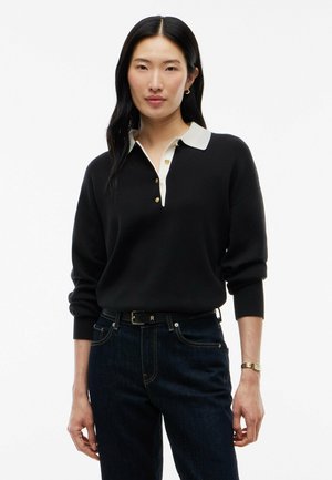 Kvinde med langt sort hår iført en sort sweater med hvid krave og knapper, mørke jeans og et armbåndsur, stående mod hvid baggrund.