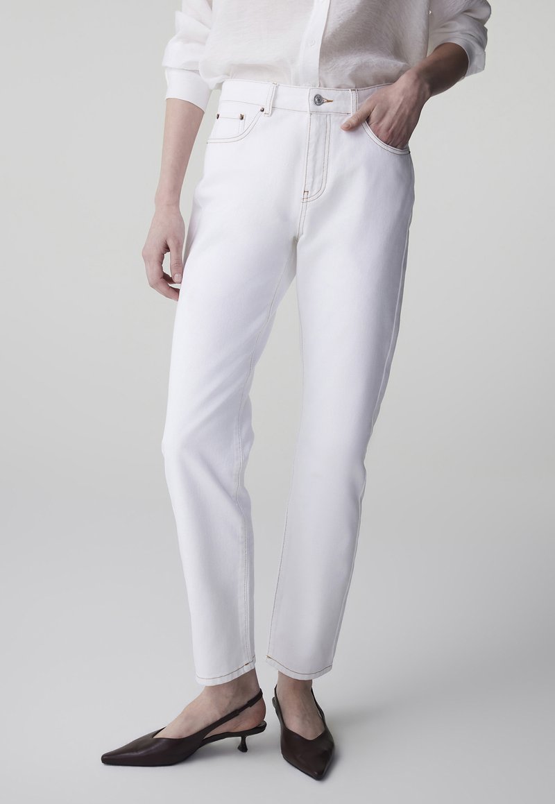 Jeans bianchi a taglio aderente con tasche frontali indossati con una camicia bianca e tacchi scuri a slingback appuntiti su sfondo chiaro.