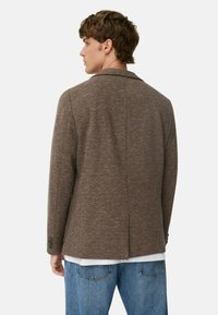 Brun, struktureret blazer med spids krave, enkelt rygventil og to-knap manchetter. Bæres over en hvid skjorte og blå jeans.