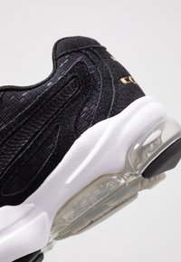 Detailní záběr na černou sportovní obuv s texturovaným svrchním materiálem, černými semišovými překryvy, bílou silnou mezipodešví a průhlednou podrážkou s vzduchovým polštářem.