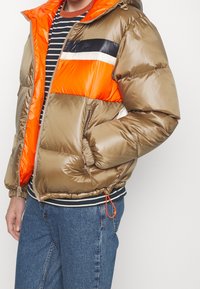 Personne portant une veste matelassée brillante couleur sable avec une doublure orange, par-dessus une chemise à rayures noires et blanches et un jean bleu, la main dans la poche.