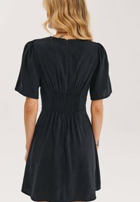 Černé šaty s krátkými nabíranými rukávy, smocked pasem a hladkým materiálem. Mají zip na zadní straně a rozšířenou sukni.