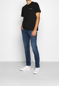 Camiseta de algodón negra con bolsillo delantero, combinada con jeans ajustados azules y zapatillas de lona blancas con acentos rojos en la suela.