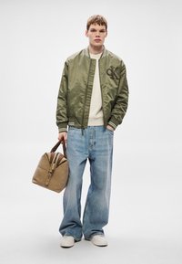 Rohekassinine bomberjope kootud kraega, valge kampsuni all, lahtised sinised teksapüksid ja pruun nahast käepidemetega kott.