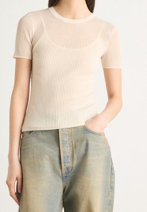 Femme portant un haut beige côtelé à manches courtes et un jean bleu délavé taille haute, une main dans la poche, sur un fond uni.
