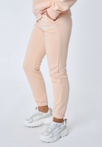 Perzikkleurige sweatpants met elastische boorden, voorzien van zijzakken en witte geborduurde tekst. Gecombineerd met chunky witte sneakers.