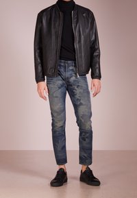 Svart läderbomberjacka, åtsittande svart polotröja, jeans i kamouflagemönster i blå denim och svarta sneakers med strukturerade ytor.