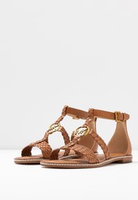 Sandales en cuir tan avec des sangles tressées, accents en logo doré, bride de cheville avec boucle et semelle plate ornée de détails en coutures blanches.