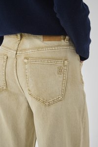 Nahaufnahme einer Person, die beige Jeans mit einer Rücktasche mit der Aufschrift "MEW" und einem dunkelblauen Pullover trägt, Hand ruht in der Seitentasche.