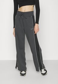 Žena v tmavě šedých širokých teplácích od Nike s bílým lemem a šňůrkou v pase, v kombinaci s černo-bílými teniskami a černým zkráceným topem.