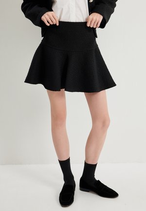 Falda negra acampanada hecha de un tejido texturizado, con una cintura ajustada y un dobladillo vaporoso. Se lleva con calcetines y zapatos negros.