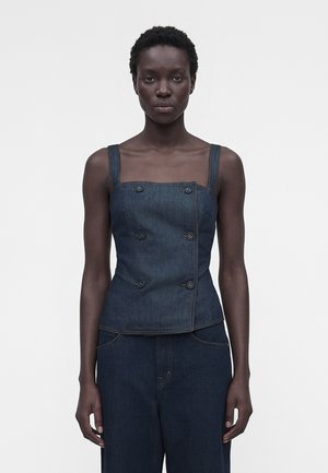 Γυναίκα που φοράει σκούρο μπλε αμάνικο διπλής σειράς τζιν τοπ και τζιν παντελόνι με φαρδιά μπατζάκια, στέκεται μπροστά σε απλό φωτεινό φόντο.