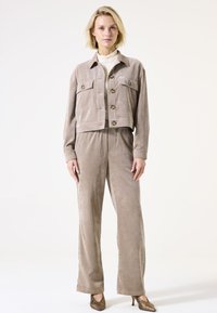 Geclipte beige corduroy jas met vier zakken, gecombineerd met bijpassende high-waisted corduroy broeken. De textuur lijkt zacht en ribbelig.