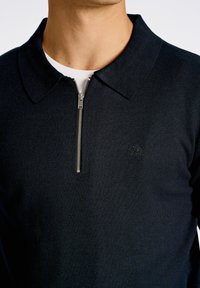 Mörkblå zip-up polo med ribbad krage och stickad textur. Silverdetalj på dragkedjan och subtil logotyp på bröstet.