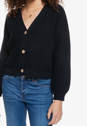 Femme portant un cardigan noir en maille avec des boutons en bois et un jean bleu, montrant la texture et les détails de la coupe du vêtement.
