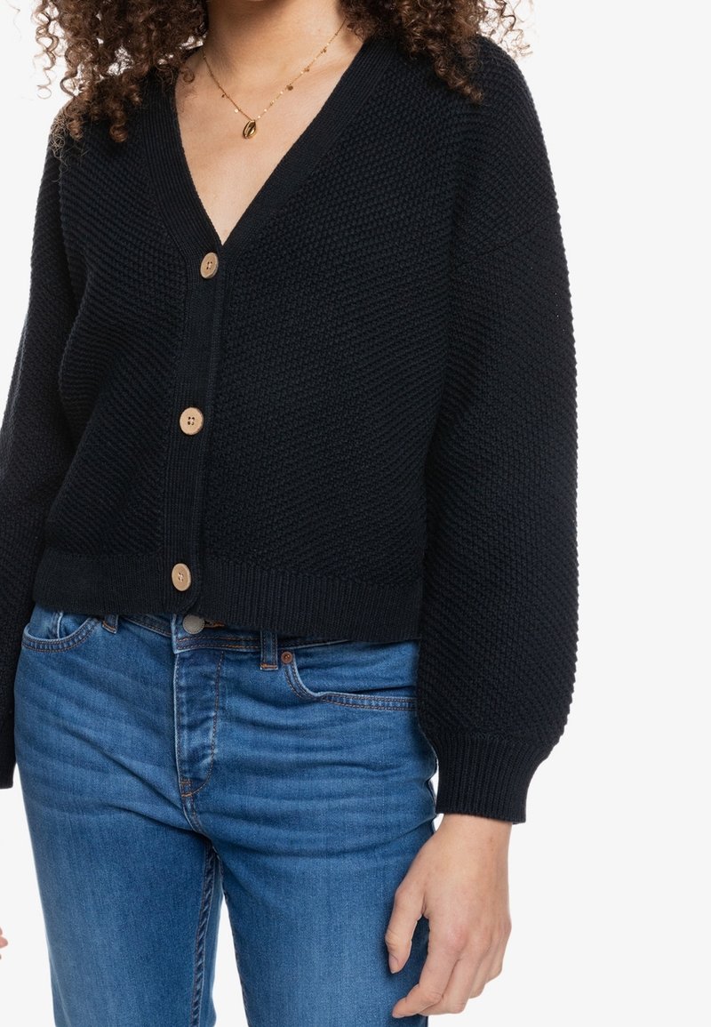 Femme portant un cardigan noir en maille avec des boutons en bois et un jean bleu, montrant la texture et les détails de la coupe du vêtement.