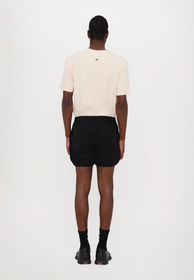 Beige kurzärmliges, strukturiertes Hemd mit Logodetail auf der Rückseite, kombiniert mit schwarzen Shorts. Sichtbare schwarze Socken und Schuhe.