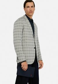 Gråternet blazer med sorte knapper, kombineret med en mørk skjorte. Bærer en mørk clutch med røde logo detaljer.