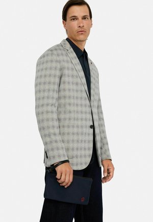 Grijze gevlokte blazer met zwarte knopen, gecombineerd met een donker shirt. Houder met een donkere clutch met rode logo details.