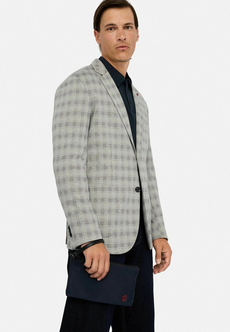 Gråternet blazer med sorte knapper, kombineret med en mørk skjorte. Bærer en mørk clutch med røde logo detaljer.