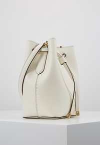 Krémová kůže kabela ve tvaru kýble s nastavitelným popruhem, stahovací šň�ůrou a zlatým kováním na bílém podstavci na jednolitém pozadí.