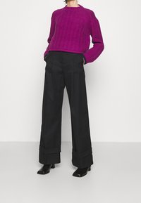 Persona vistiendo un suéter de punto trenzado magenta, pantalones anchos de cintura alta negros con dobladillos, y botines negros de tacón, de pie con las manos en los bolsillos.