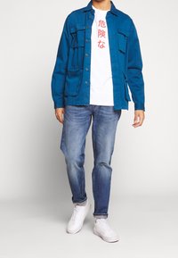 Muž v modré bunda na knoflíky přes bílé tričko s červenými asijskými znaky, modrých džínách a bílých teniskách stojící na jednobarevném pozadí.