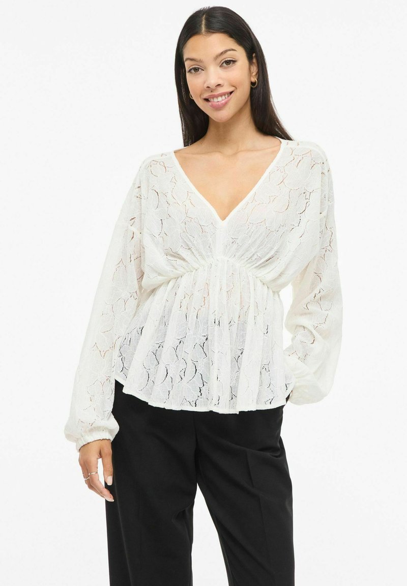 Biała koronkowa bluzka z długimi, luźnymi rękawami, głębokim dekoltem w serek i marszczoną talią. Przezroczysta tkanina z delikatnym kwiatowym wzorem. Stylizowana z czarnymi spodniami.