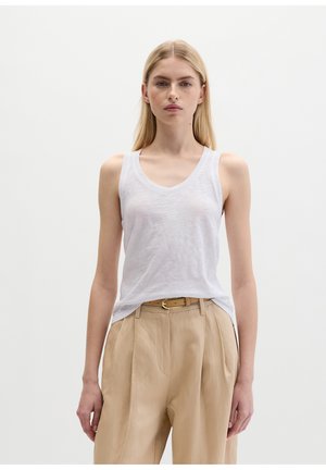 Blonde woman wearing a white sleeveless V-neck top tucked into beige high-waist pleated trousers with a tan belt, standing against a white background.