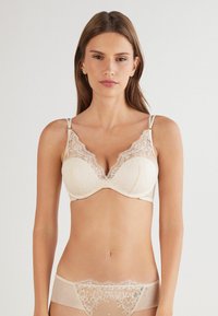Sostén de color beige con aros, detalles de encaje floral, bordes ondulados y tirantes ajustables; combinado con unas braguitas a juego con borde de encaje.