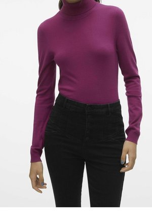 Mujer con un suéter de cuello alto ajustado de color púrpura metido dentro de pantalones negros de cintura alta con botones, de pie contra un fondo liso.